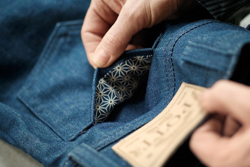 Eine Hand öffnet die Tasche einer Jeans und zeigt das gemusterte Innenfutter, daneben das Lederlabel von POTT.