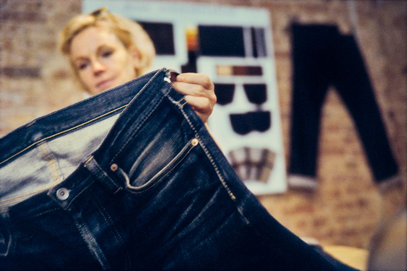 Manuela Pott hält den Bund einer dunklen Jeans hoch. Im Hintergrund eine Musterkarte mit Taschen- und Stoffproben an einer Backsteinwand.