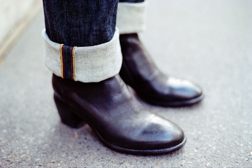 Nahaufnahme eines umgeschlagenen Jeanssaums mit farbiger Webkante über braunen Lederstiefeletten.