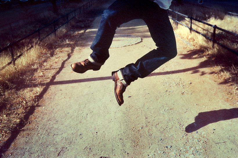 Eine staubige Straße von oben, ins Bild ragen die Beine einer Person, die im Luftsprung die Hacken aneinanderschlägt. Sie trägt eine Jeans von POTT.